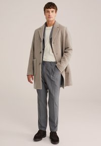 Man in een beige overjas, witte crewneck shirt, grijs gestreepte broek en donkerbruine loafers, staand met één hand in zijn zak.