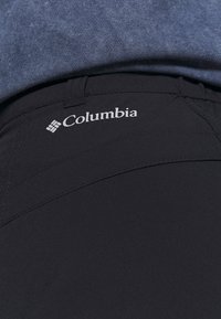 Musta Columbia-housut ovat valmistettu sileästä, kevyestä kankaasta, jossa on hienostunut kiilto. Vyötärö on suunniteltu mukavaksi, ja siinä on pieni logo printattuna.