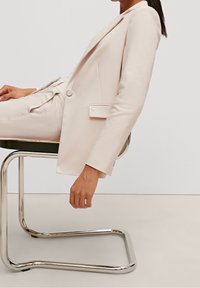 Jasno-beżowa marynarka o dopasowanym kroju, z pojedynczym zapięciem na guzik i gładką teksturą, noszona na pasujących spodniach, podczas siedzenia na metalowym krześle.