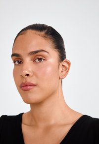 Zlatne hoop naušnice s tankim, izduženim elementom. Glatka tekstura; mali, okrugli detalj na uhu. Model prikazuje naušnice.