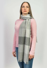 Sciarpa di lana grigia e bianca a quadri con bordi frangiati, appesa casualmente attorno al collo; indossata sopra un maglione rosa e jeans blu.