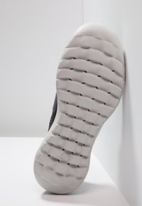Semelle de chaussure en caoutchouc gris texturé, avec un motif de bande de roulement et un bout arrondi. Incliné contre un fond clair.