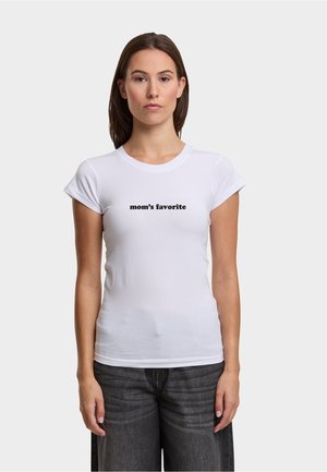 Kobieta stojąca przodem, ubrana w dopasowany biały t-shirt z napisem "ulubiona mama" w czarnym kolorze oraz ciemnoszare dżinsy.