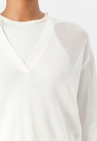 Hvit mesh langermet topp med V-hals og ribbet indre lag. Har en løs passform og teksturert, pustende materiale med rutenettmønster.