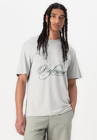 Lys grå bomulds T-shirt med korte ærmer, prydet med grøn tekst og emblem på forsiden, afslappet pasform og en glat tekstur.