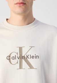 Svjetlo bež dukserica s rebrastim okovratnikom. Na prednjoj strani nalazi se natpis "CK" i "Calvin Klein" u kontrastnoj bež vezici. Glatka textura.
