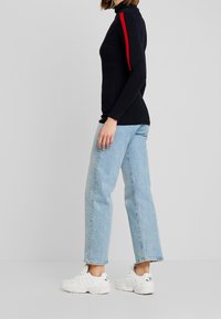 Marineblauwe coltrui met rode streep op de schouder, ribbel structuur, gecombineerd met lichtblauwe straight-leg jeans en witte sportschoenen.