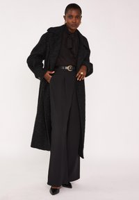 Manteau long noir à finition texturée, porté sur un chemisier noir et un pantalon à jambes larges. Comprend une ceinture cintrée et des escarpins à bout pointu.