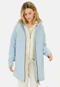 Lichtblauwe waterdichte jas met een ritssluiting, verstelbare capuchon, elastische manchetten en pocketdetails, gedragen over een crèmekleurige top en broek.