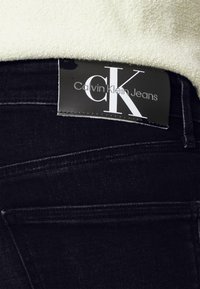 Jeans de denim negros con un parche de logo de Calvin Klein Jeans negro cerca de la cintura, combinados con una blusa blanca texturada.