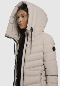 Veste beigne rembourrée avec une grande capuche, zip noir et écussons. L'extérieur est lisse ; la doublure intérieure a une texture brillante.