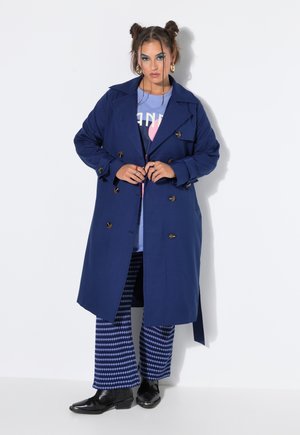 Donna con acconciatura a doppio chignon che indossa un trench blu, pantaloni blu fantasia e scarpe nere, in piedi davanti a uno sfondo bianco.