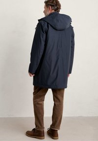 Abrigo azul marino, hasta la cadera, con capucha. Tejido liso, resistente al agua, con bolsillos laterales y corte recto. Llevar encima de pantalones de pana marrón.
