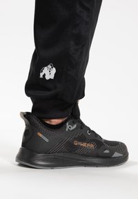 Zwart sportieve schoen met een gestructureerde mesh bovenkant, grijze accenten, rubbere zool en een oranje logo. Gecombineerd met zwarte nauwsluitende joggers met een gorilla logo.