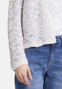 Stickad cardigan med ett vågmönster i pastellfärger, med openwork-detaljer och ribbad mudd, tillsammans med blå denimjeans.
