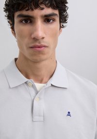 Camisa polo gris claro con un cuello clásico, dos botones y un logo bordado de una calavera y huesos azules en el lado izquierdo del pecho.
