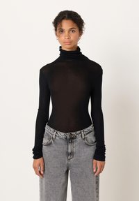 Noor naine mustas pika varrukaga kõrge kaelusega pluusis ja helehallides kõrge vöökohaga teksapükstes, seistes lihtsa tausta ees.