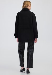 Cappotto di lana nera con una vestibilità su misura, ampio colletto e dettaglio con cintura sul retro. Il modello indossa pantaloni in pelle neri e tacchi a punta.