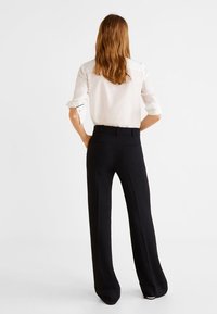 Chemise blanche à manches longues et pantalons noirs amples. La chemise a les manches retroussées ; les pantalons sont en tissu lisse et ont des jambes droites.