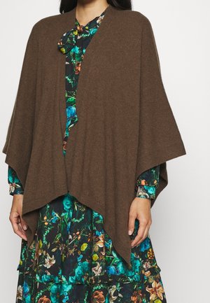 Vrouw draagt een bruine gebreide poncho over een langarmige donkere bloemenjurk met groene, blauwe en oranje bloemen op een witte achtergrond.