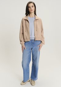 Giacca beige con zip e cordini, top azzurro chiaro e jeans a gamba larga azzurri chiari. Calzature: mocassini beige con dettagli in metallo.