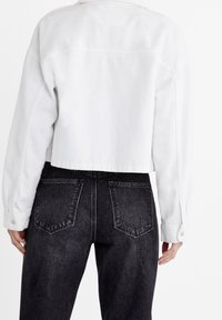 Chaqueta de mezclilla blanca recortada con un cierre de botones en la parte delantera y dos bolsillos laterales, combinada con unos jeans oscuros de cintura alta en gris que tienen bolsillos traseros.