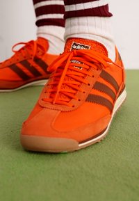 Narančaste sportske cipele od suede-a i mrežaste tkanine s smešnim kožnim detaljima, koje imaju vezice, gumene potplate i klasični Adidas logo na jeziku.