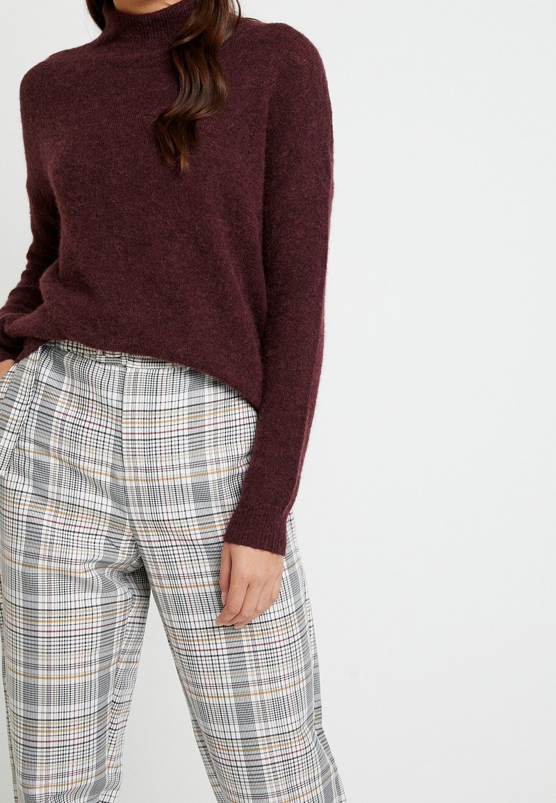 Suéter de punto burdeos con un cuello alto, combinado con pantalones de cuadros que presentan líneas grises, blancas y de colores. Se observa una textura suave.