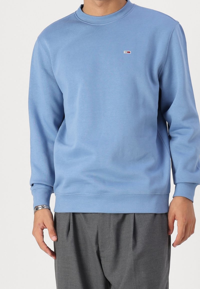 Homme portant un sweat-shirt bleu clair à manches longues et un pantalon gris, avec un petit logo rectangulaire sur la poitrine gauche.