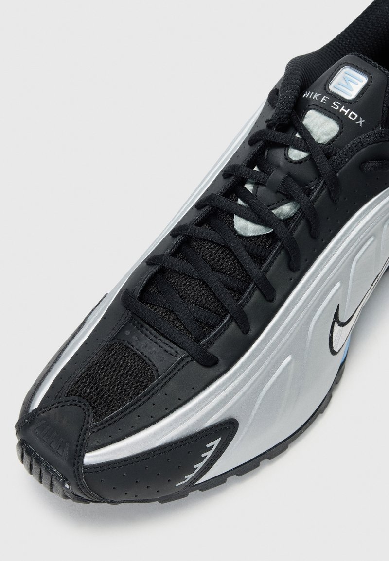 Zapatilla Nike Shox negra y plateada, fabricada con materiales de malla y sintéticos, que cuenta con un cuello acolchado, punta redondeada y suela texturizada.