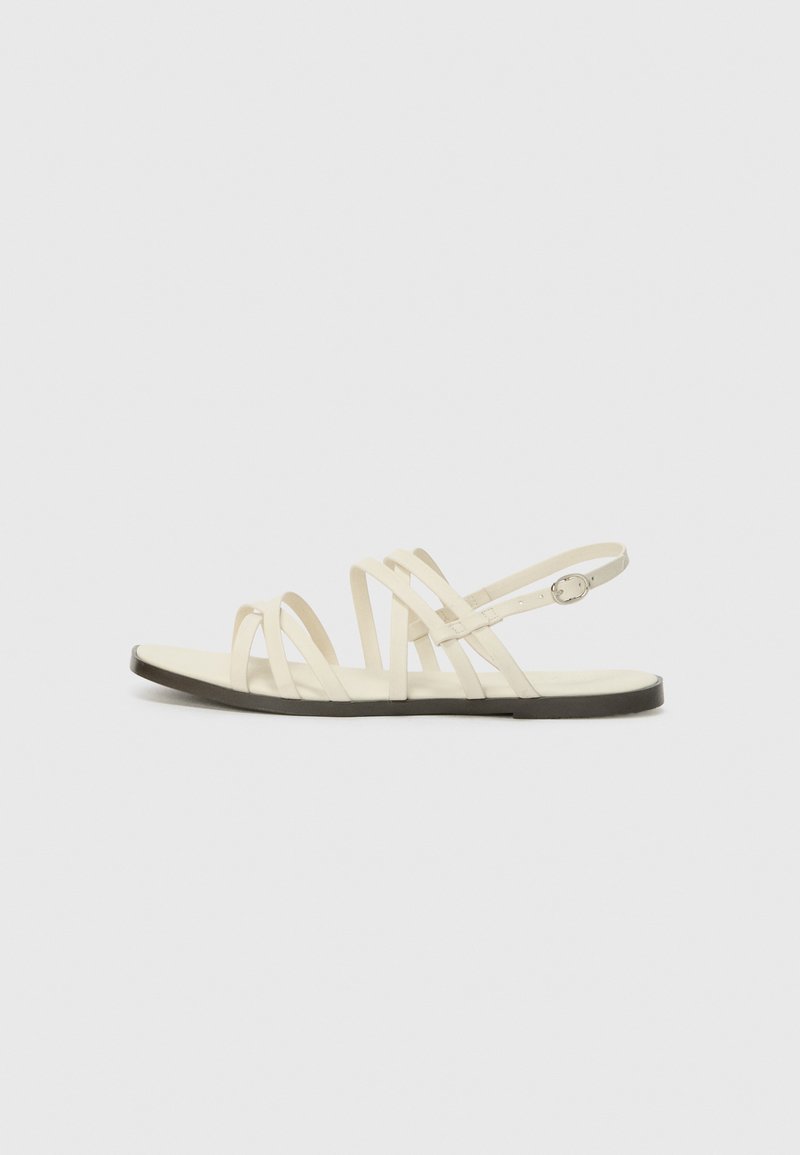 Strappy sandaler i kremfarge laget av syntetisk materiale, med flere smale stropper og en flat brun såle med justerbar ankelreim.