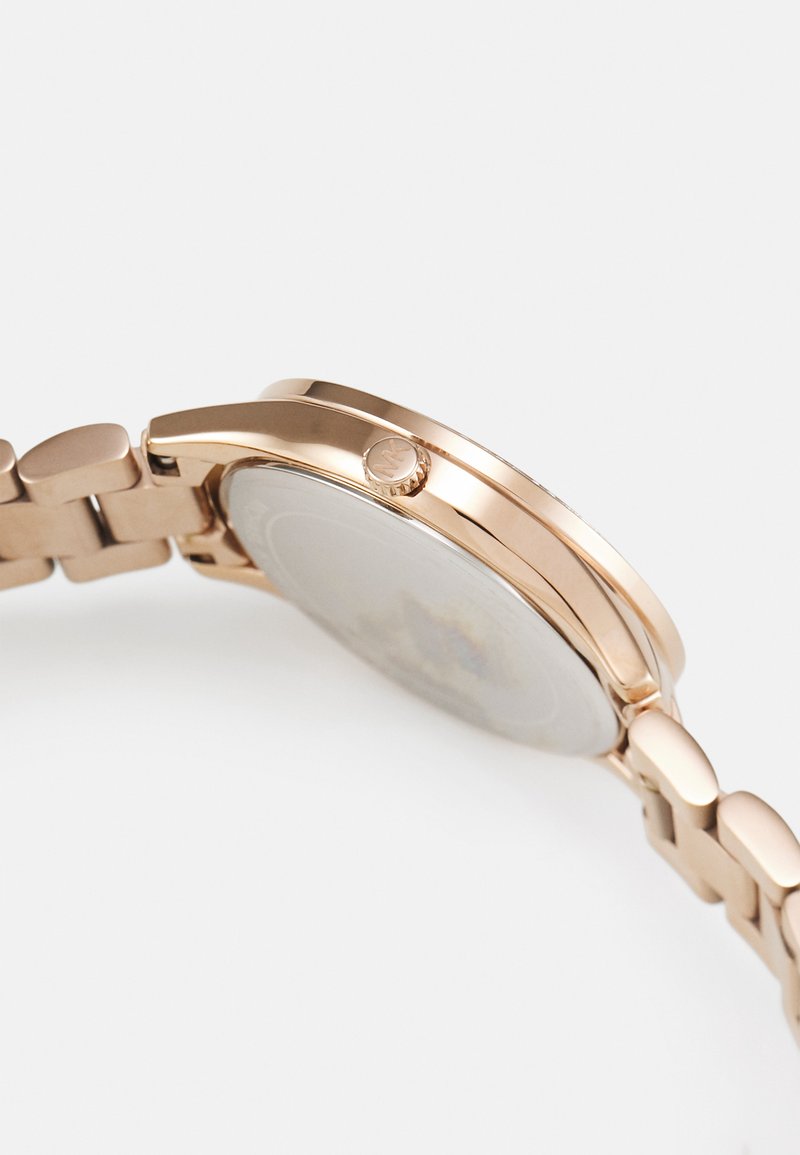 Różowo-złoty metalowy zegarek na rękę pokazany z boku, z podkreśloną koroną z wytłoczonym logo i połączoną bransoletą na białym tle.