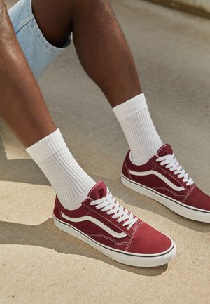 Person iført hvide ribstrikkede crew-sokker og røde lave sneakers, der sidder på en beige betonoverflade med benene delvist bøjede.