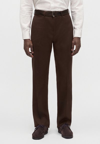 Homme portant un pantalon de costume marron foncé, une ceinture noire, des chaussures marron foncé et une chemise blanche boutonnée, debout devant un fond uni.