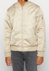 Giacca bomber beige in tessuto satin, con zip frontale, polsini a coste e tasche laterali. Include una tasca con zip sulla manica.