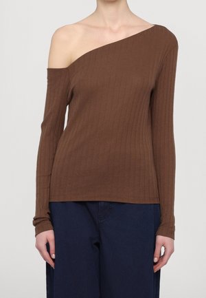 Femme portant un haut marron côtelé une épaule à manches longues, associé à un pantalon bleu foncé sur un fond uni.