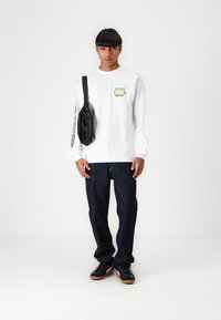 Langærmet hvid skjorte med grøn grafik, sort crossbody taske, mørkeblå jeans og sorte sneakers med gummisål. Enkel stil, minimalistisk design.