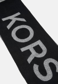 Černý šátek s velkými kontrastními šedými písmeny, která vypisují "KORS." Vyrobeno z jemné látky se mírně chlupatou texturou.