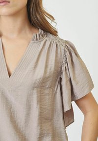 Top beige chiaro con scollatura a V, maniche corte a volant e dettagli arricciati sulle spalle. Realizzato in un tessuto texturizzato.