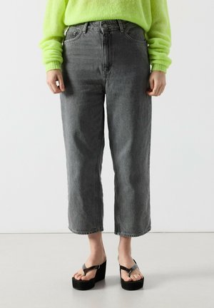 Personne portant un jean gris court taille haute, un pull fluo vert en matière pelucheuse et des sandales plateformes noires à claquettes, debout sur un sol clair.