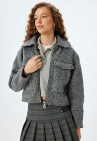 Giacca grigia, strutturata e corta con chiusura con zip e tasca sul petto con pattina, abbinata a un polo grigio e una gonna a quadretti. Orecchini circolari visibili.