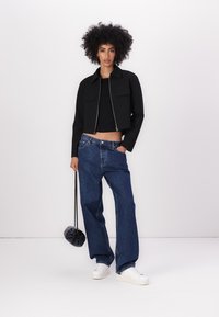 Zwarte cropped jas met ritssluiting, twee voorzakken; gecombineerd met losse blauwe denim jeans en witte sneakers; houdt een kleine zwarte tas vast.
