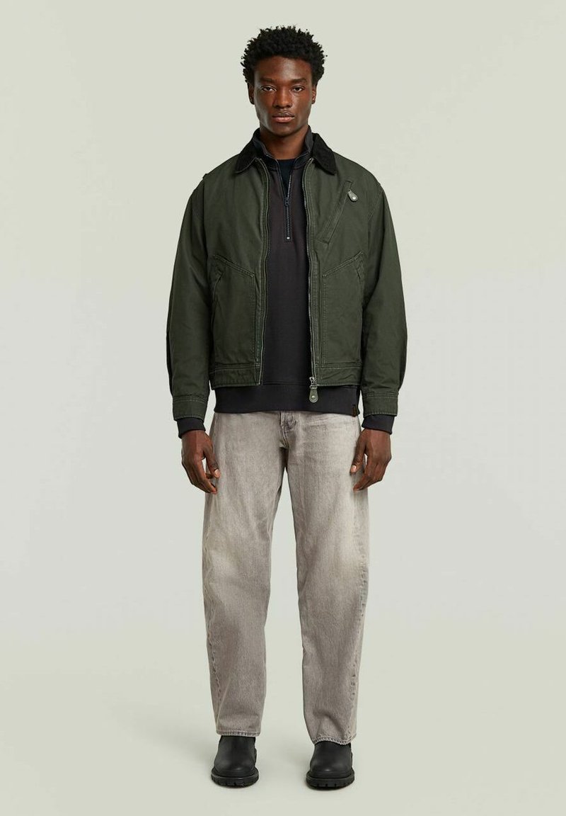 Veste bomber vert olive avec un col noir, fermeture éclair à l'avant et deux poches latérales, portée par-dessus un haut noir et un jean gris clair, assortie avec des bottes noires.