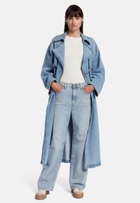 Un trench-coat en denim bleu clair sur un haut blanc, associé à un jean large bleu clair et des sneakers crème avec des semelles brunes. Boucles d'oreilles en argent.