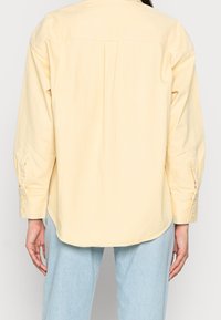 Personne portant une chemise beige à manches longues et un jean bleu clair, vue de dos, les bras détendus le long du corps.