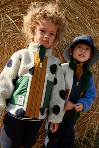 To børn bærer farverige jakker: den ene i hvid fleece med sorte prikker, grønne og brune detaljer; den anden i blå og grøn tekstureret fleece.