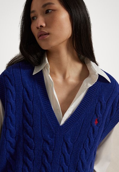 Blue knit vest with cable pattern and V-neck design, layered over a white collared shirt. Features a small red logo on the chest.
