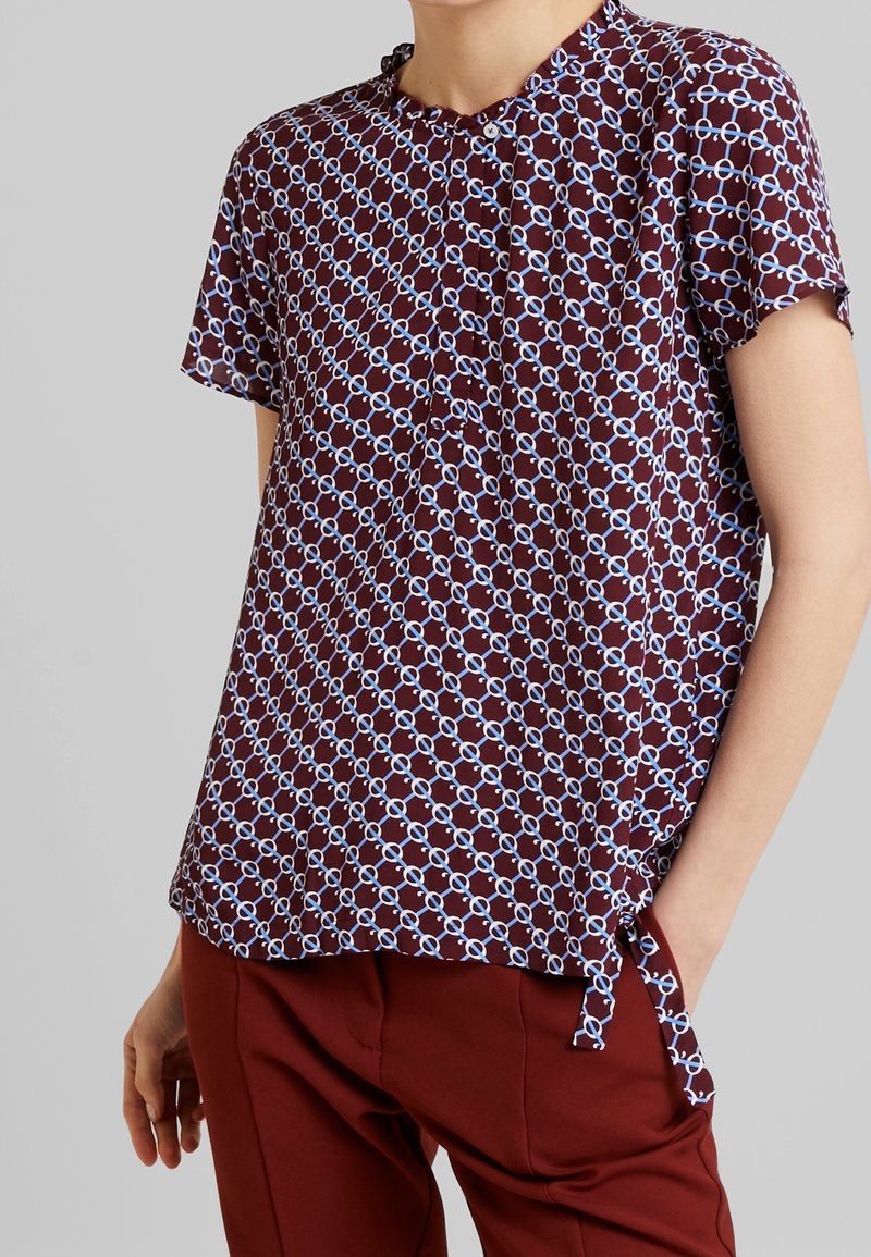 Blouse bordeaux à manches courtes avec motif chaîne géométrique blanc et bleu, associée à un pantalon bordeaux, portée par une personne debout devant un fond gris.