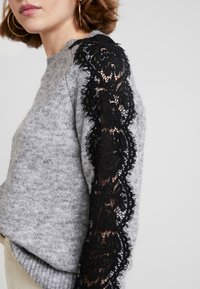 Donna che indossa un maglione grigio a maglia con pannello in pizzo floreale nero che scende lungo la manica esterna, abbinato a orecchini a cerchio dorati.