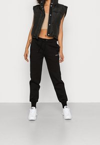 Czarna kamizelka puchowa z zatrzaskami, beżowy krótki top, czarne dresy z troczkiem i białe sneakersy. Prosty design, sportowy wygląd.